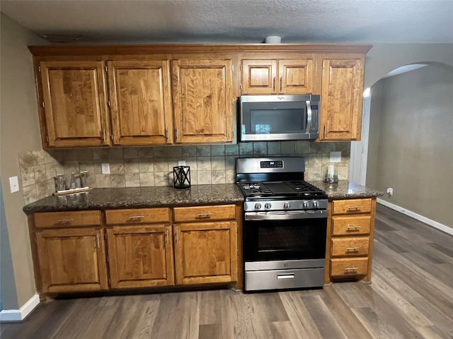 kitchen with appliances with stainless steel finishes, dark stone countertops, dark hardwood / wood-style floors, and tasteful backsplash