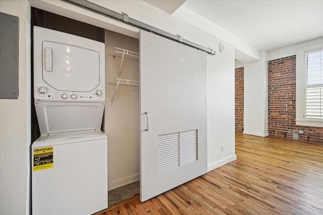 washroom featuring wood-type flooring, stacked washer and dryer, electric panel, a barn door, and brick wall