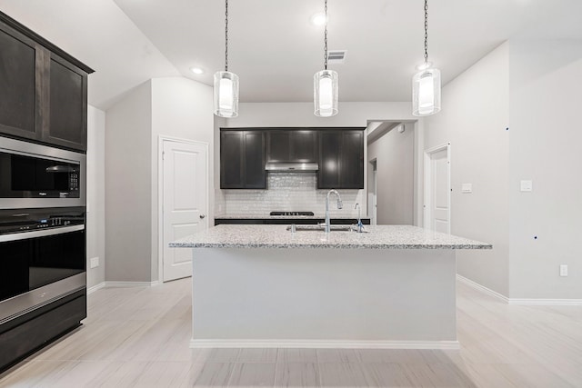 kitchen with tasteful backsplash, a kitchen island with sink, a sink, light stone countertops, and built in microwave