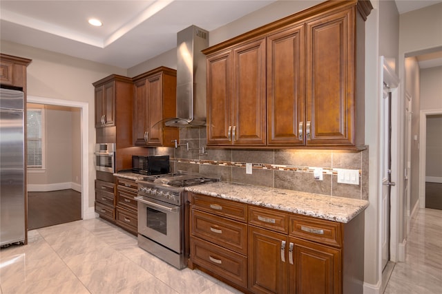 kitchen with backsplash, high end appliances, light stone countertops, a tray ceiling, and wall chimney range hood