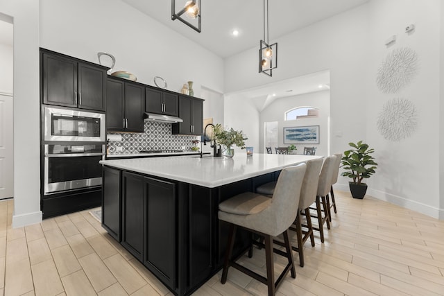 kitchen with a kitchen island with sink, tasteful backsplash, stainless steel microwave, light hardwood / wood-style flooring, and decorative light fixtures