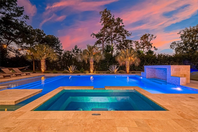 pool at dusk with an in ground hot tub, pool water feature, and a patio area