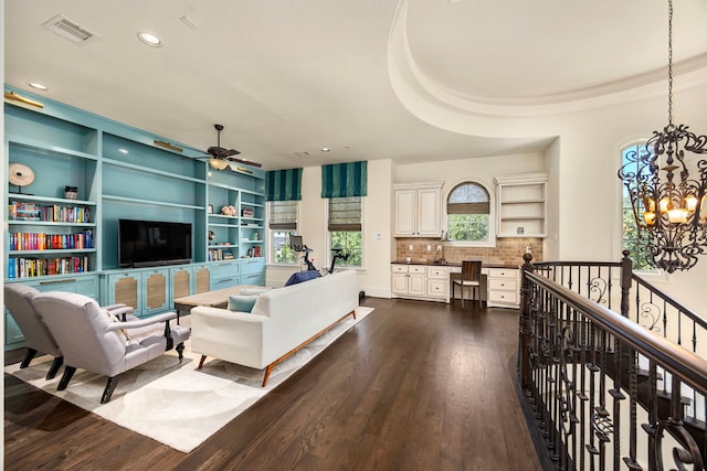 living room featuring built in desk, dark hardwood / wood-style floors, ceiling fan with notable chandelier, built in shelves, and a raised ceiling