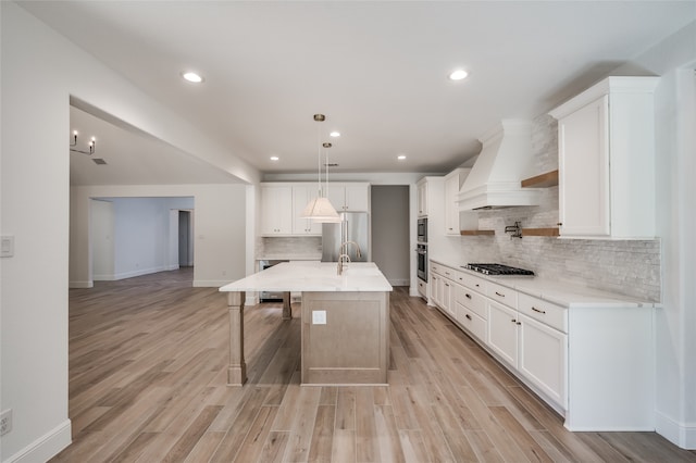 kitchen with premium range hood, a center island with sink, light hardwood / wood-style flooring, appliances with stainless steel finishes, and white cabinetry