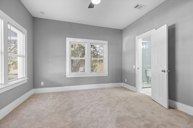 unfurnished bedroom featuring light colored carpet, ensuite bath, multiple windows, and ceiling fan