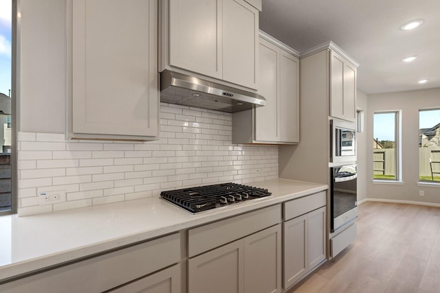 kitchen with stainless steel appliances, light countertops, light wood-type flooring, under cabinet range hood, and backsplash