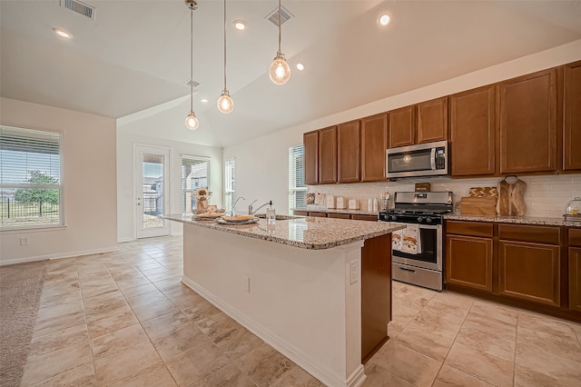 kitchen featuring stainless steel appliances, decorative light fixtures, light stone countertops, decorative backsplash, and an island with sink