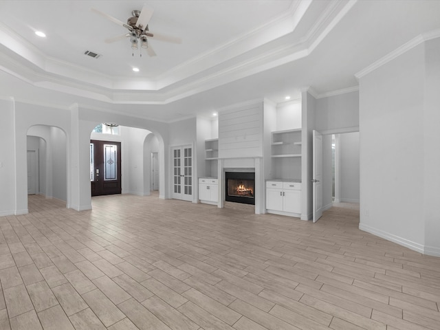 unfurnished living room with a raised ceiling, built in shelves, ceiling fan, ornamental molding, and light wood-type flooring