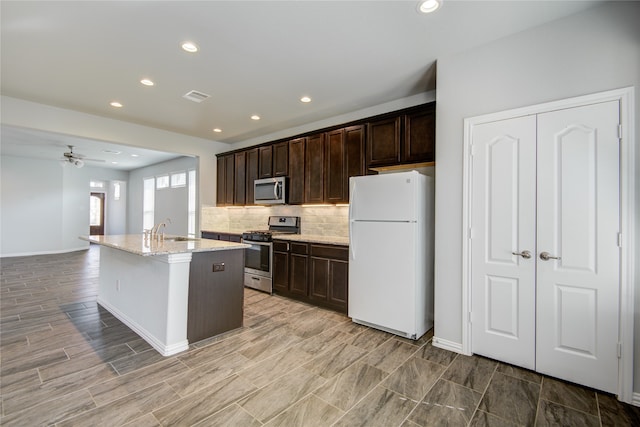 kitchen with light stone countertops, appliances with stainless steel finishes, a kitchen island with sink, ceiling fan, and sink