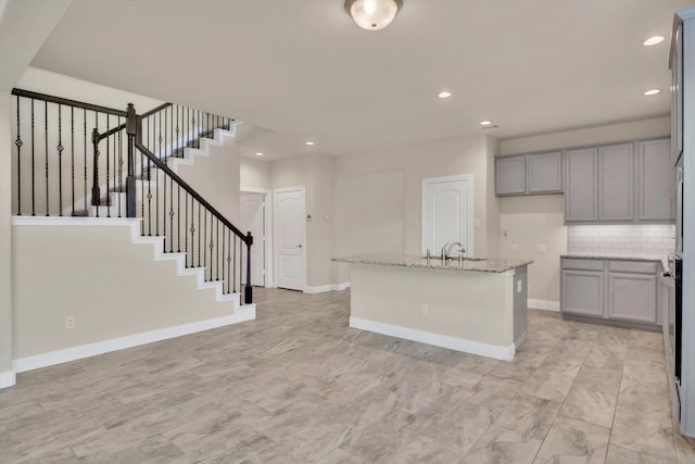 kitchen featuring sink, gray cabinetry, a center island with sink, light stone countertops, and decorative backsplash