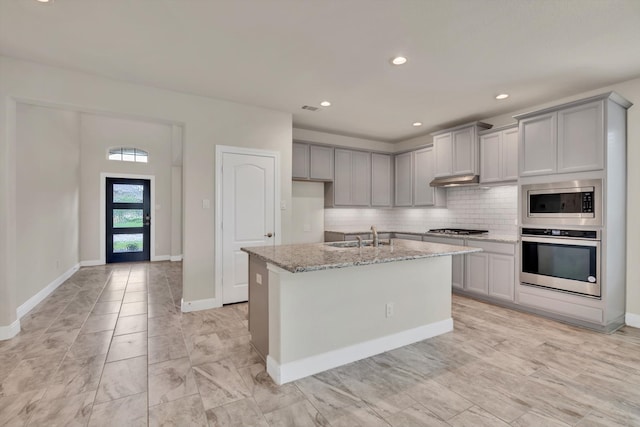 kitchen with light stone counters, gray cabinets, stainless steel appliances, a kitchen island with sink, and sink