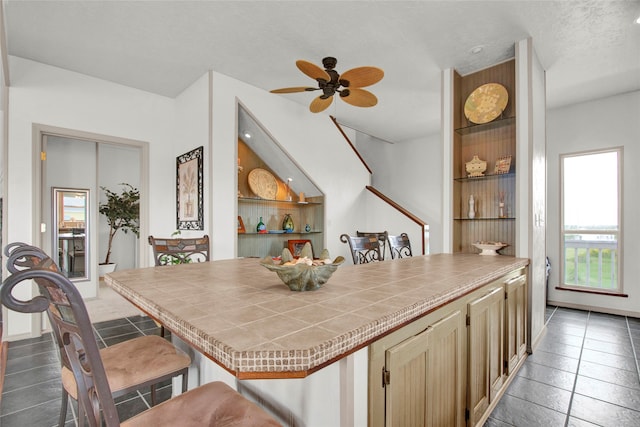 kitchen featuring ceiling fan, dark tile patterned flooring, a textured ceiling, tile countertops, and a breakfast bar area