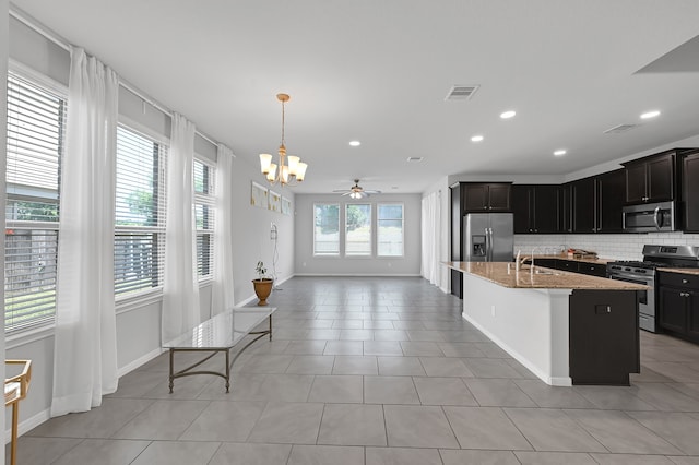 kitchen with light stone counters, ceiling fan with notable chandelier, decorative backsplash, a center island with sink, and appliances with stainless steel finishes