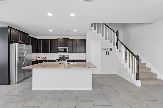 kitchen featuring a center island with sink, dark brown cabinets, stainless steel appliances, and light stone counters