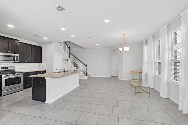 kitchen with an island with sink, an inviting chandelier, stainless steel appliances, decorative backsplash, and light stone countertops