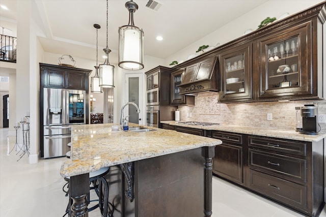 kitchen with appliances with stainless steel finishes, premium range hood, dark brown cabinetry, a kitchen island with sink, and sink