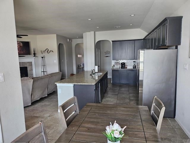 kitchen with light stone counters, sink, a tiled fireplace, stainless steel refrigerator, and an island with sink