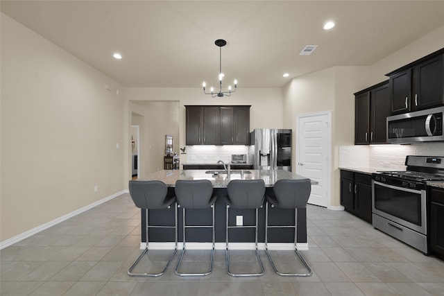 kitchen featuring light stone counters, a kitchen island with sink, stainless steel appliances, a notable chandelier, and backsplash