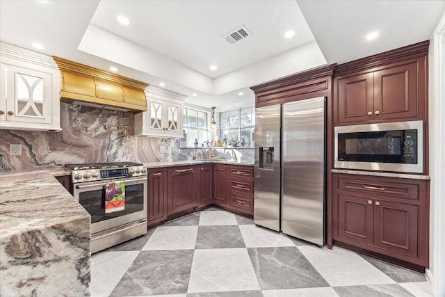 kitchen with light stone counters, premium range hood, sink, built in appliances, and backsplash