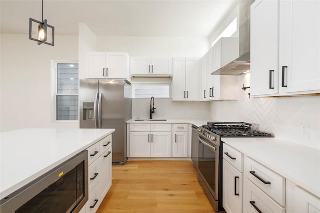 kitchen featuring white cabinets, wall chimney range hood, light wood-type flooring, decorative light fixtures, and stainless steel appliances