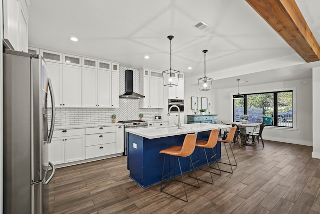 kitchen featuring stainless steel appliances, wall chimney range hood, pendant lighting, white cabinetry, and an island with sink