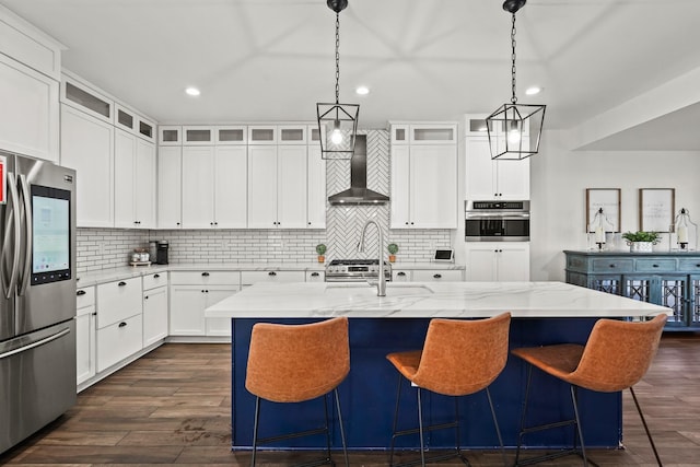 kitchen with stainless steel appliances, wall chimney range hood, light stone counters, a center island with sink, and white cabinets