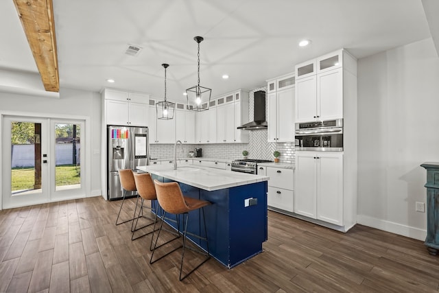 kitchen with a center island with sink, wall chimney range hood, beam ceiling, white cabinetry, and stainless steel appliances