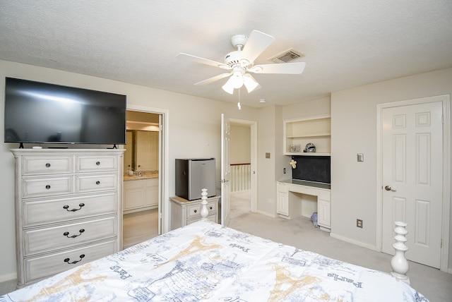 bedroom featuring built in desk, ceiling fan, a textured ceiling, ensuite bathroom, and light colored carpet