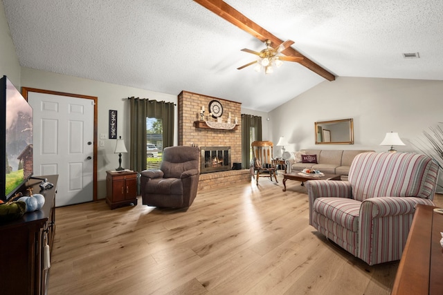 living room featuring a brick fireplace, light wood-type flooring, ceiling fan, vaulted ceiling with beams, and a textured ceiling