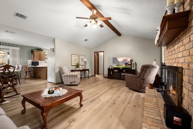 living room with a brick fireplace, light hardwood / wood-style floors, ceiling fan, vaulted ceiling with beams, and a textured ceiling