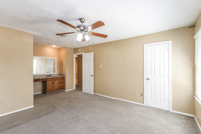 unfurnished bedroom featuring a textured ceiling, ceiling fan, built in desk, ensuite bath, and light colored carpet