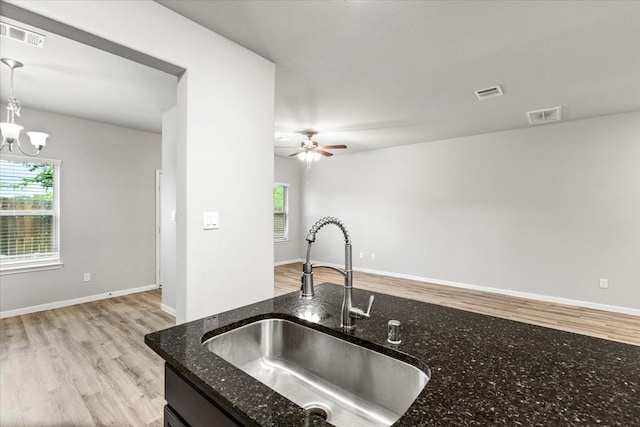 kitchen with sink, ceiling fan with notable chandelier, dark stone counters, hanging light fixtures, and light hardwood / wood-style flooring