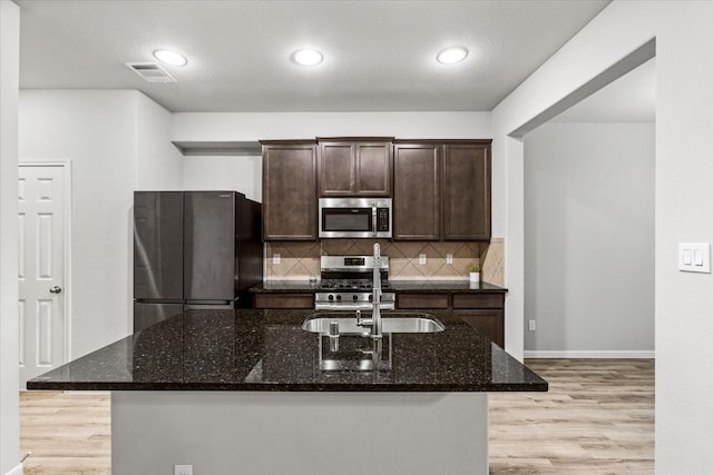 kitchen with dark brown cabinetry, backsplash, dark stone counters, appliances with stainless steel finishes, and light wood-type flooring