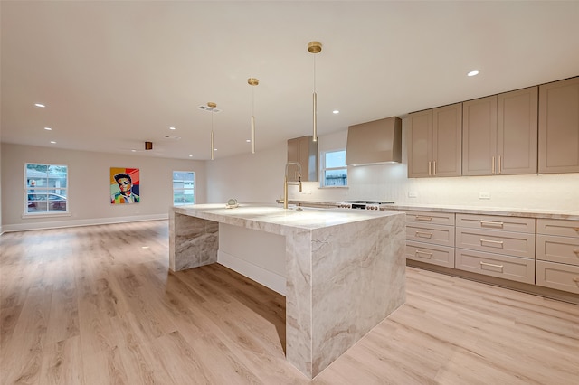 kitchen with wall chimney exhaust hood, an island with sink, hanging light fixtures, light hardwood / wood-style flooring, and light stone countertops