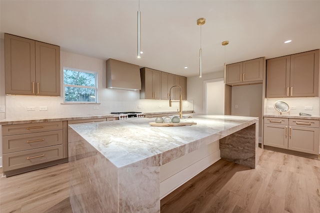 kitchen with wall chimney range hood, a kitchen island with sink, light stone countertops, light wood-type flooring, and pendant lighting