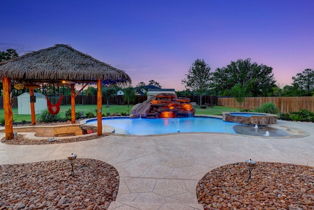 pool at dusk featuring an in ground hot tub, a storage shed, a water slide, pool water feature, and a patio area