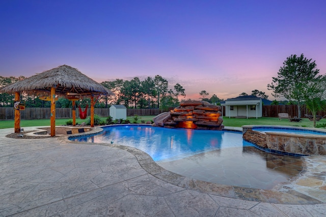 pool at dusk with an in ground hot tub, a patio area, a fire pit, and a storage unit