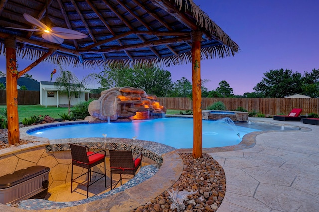 pool at dusk with a gazebo, ceiling fan, pool water feature, a patio, and an in ground hot tub