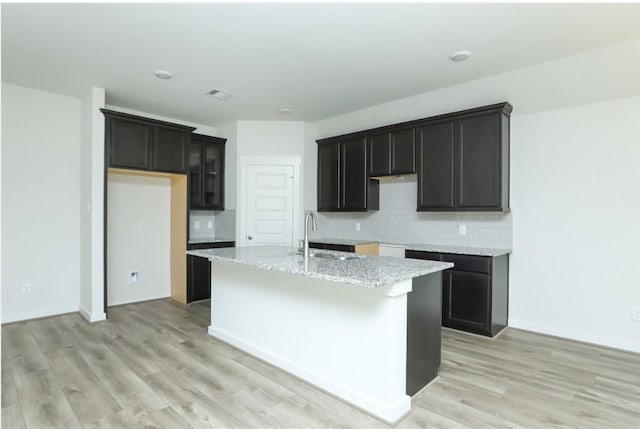 kitchen featuring light wood-type flooring, light stone countertops, a kitchen island with sink, and sink