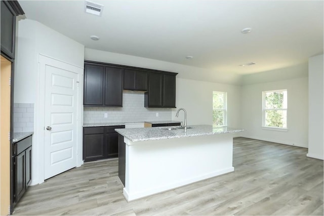 kitchen with light hardwood / wood-style floors, a center island with sink, sink, light stone countertops, and backsplash