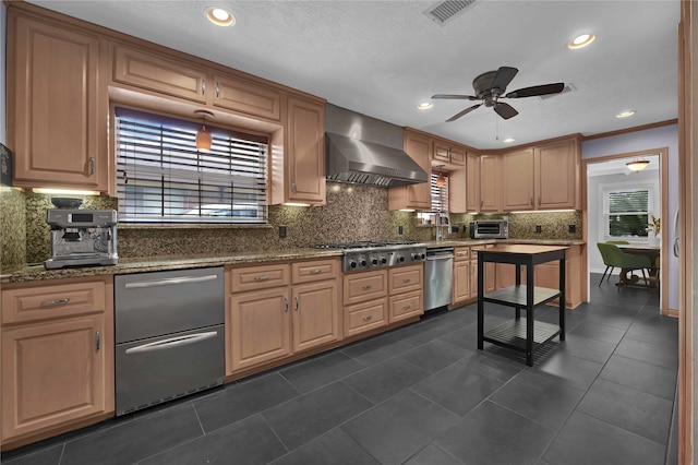 kitchen with ceiling fan, dark tile patterned flooring, wall chimney exhaust hood, appliances with stainless steel finishes, and dark stone countertops