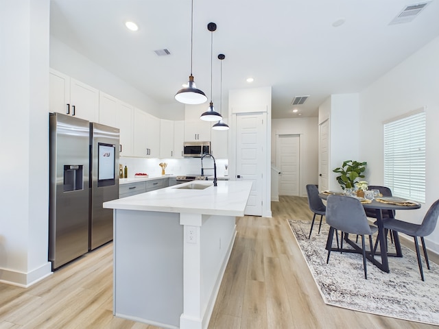 kitchen with decorative light fixtures, a center island with sink, white cabinetry, stainless steel appliances, and light wood-type flooring