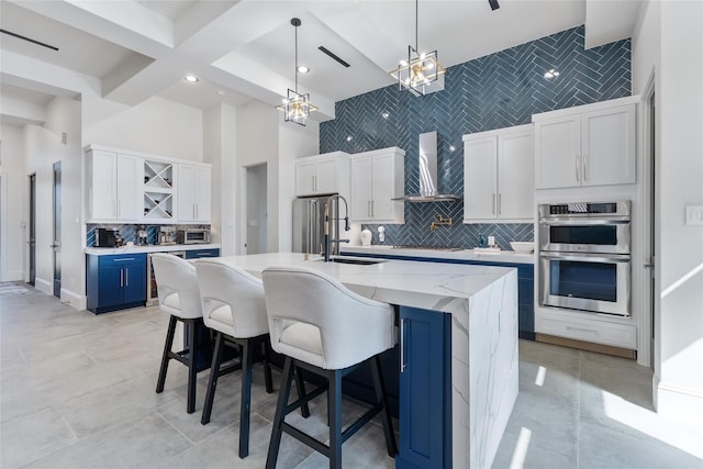 kitchen with white cabinets, wall chimney exhaust hood, a large island with sink, blue cabinetry, and a towering ceiling