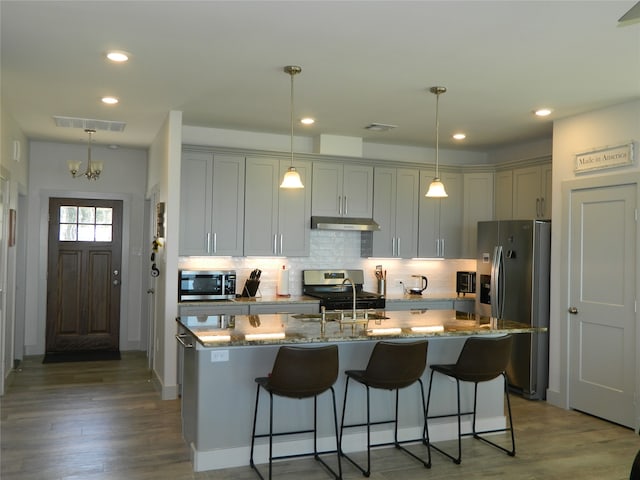 kitchen with light stone counters, decorative light fixtures, a kitchen island with sink, dark wood-type flooring, and appliances with stainless steel finishes