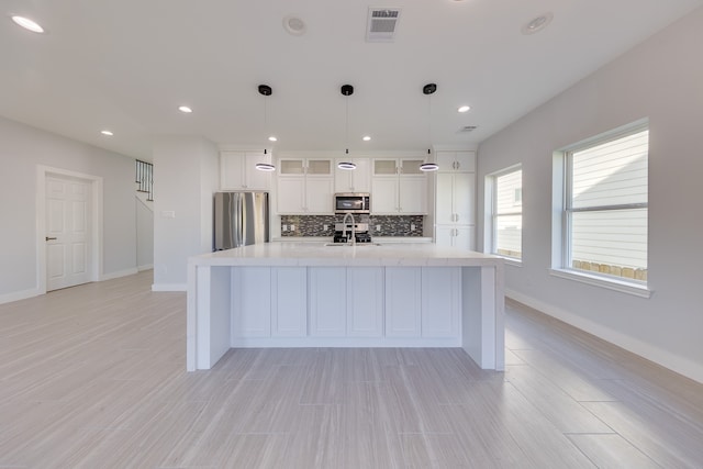 kitchen with decorative light fixtures, a center island with sink, backsplash, white cabinetry, and stainless steel appliances