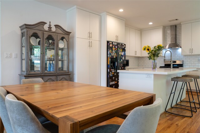 kitchen with tasteful backsplash, wall chimney exhaust hood, black fridge, white cabinetry, and light hardwood / wood-style flooring