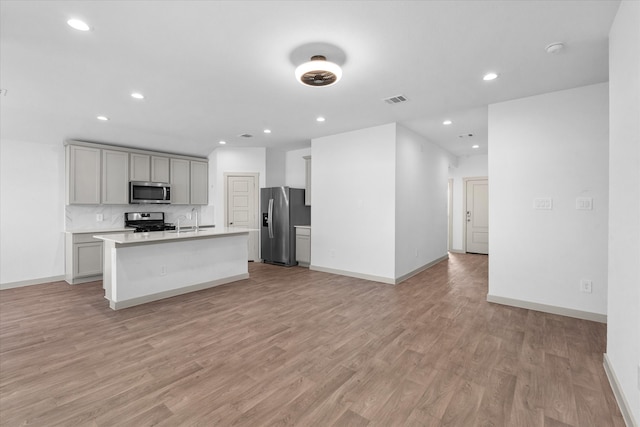 kitchen with a kitchen island with sink, gray cabinetry, appliances with stainless steel finishes, light hardwood / wood-style floors, and tasteful backsplash