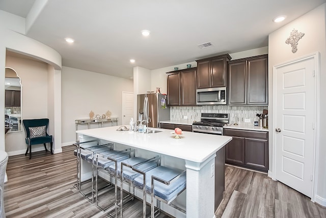 kitchen with decorative backsplash, hardwood / wood-style floors, dark brown cabinets, an island with sink, and stainless steel appliances