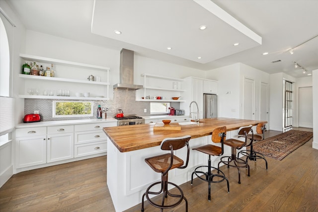 kitchen with white cabinets, wooden counters, a center island with sink, dark wood-type flooring, and wall chimney range hood