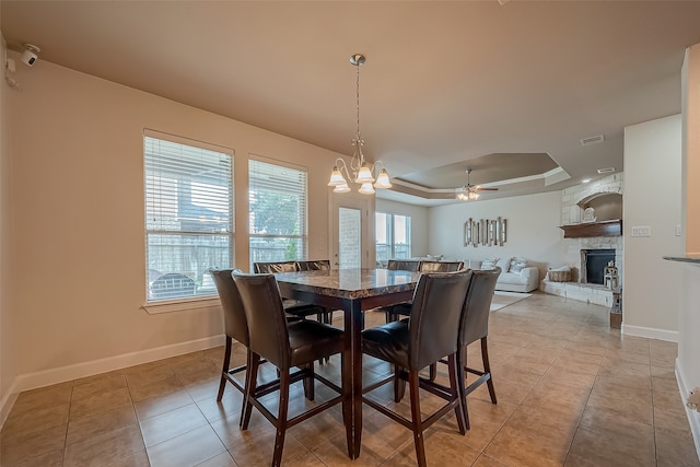 tiled dining room with ceiling fan with notable chandelier, a stone fireplace, and a raised ceiling